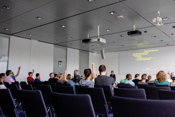 A lecture room with a group of people seen from behind. They look towards a projected slide at the front right of the frame. Many of them have their hands raised to indicate they want to ask something.