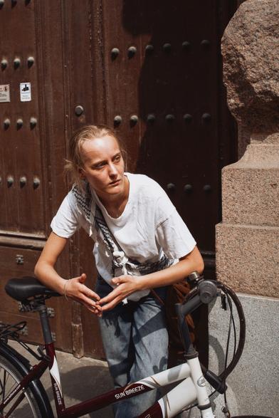 A women leaning on her bike, she has white tshirt and a keffiyeh on her shoulder.