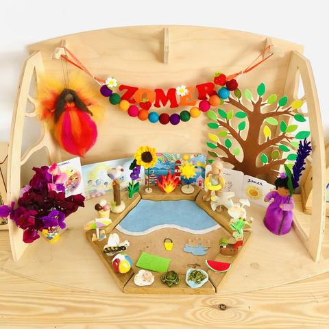 A square photo of our seasonal table; a wooden stage with on it a wooden seasonal ring with Summer themed pegs like a sunflower, watermelon and a beach ball and in the middle a homemade flat felt beach and sea. At the back are two small buntings, one is a string of colourful felt balls and the other is in red and orange felt the word “Zomer” which is Dutch for Summer, decorated with strawberries and strawberry flowers. The hanging felt fairy of this season has black skin, brown long hair, a dress in orange, red and pink, and wings in yellow and red. The wooden seasonal tree that had pink blossoms before, is now filled with green leaves. To the left of the ring is a little yellow vase filled with magenta, pink and purple sweet pea flowers. To the right of the ring is a felt doll with brown skin, black curls and a purple dress, holding a lavender branch bigger than her.