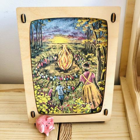 A wooden lamp with an A6 sized postcard showing a chalkboard drawing of a big group of people wearing flower crowns around a huge fire in a field against the setting sun
