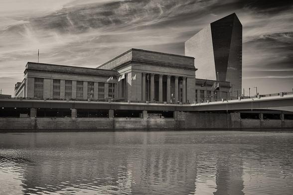 A large neoclassical train station across a river. To the right of the station is a modernist glass office tower.