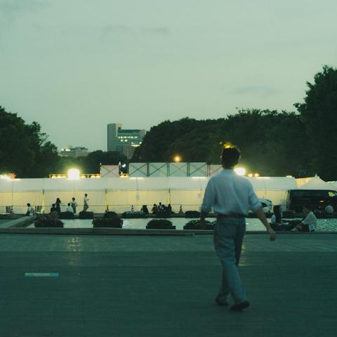A person walks toward an illuminated area at dusk, surrounded by white tents and a row of small bushes. People gather near a fountain in the background. Trees and a cityscape with tall buildings are visible in the distance under a dimly lit sky.