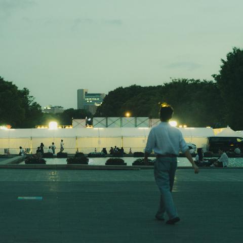 A person walks toward an illuminated area at dusk, surrounded by white tents and a row of small bushes. People gather near a fountain in the background. Trees and a cityscape with tall buildings are visible in the distance under a dimly lit sky.