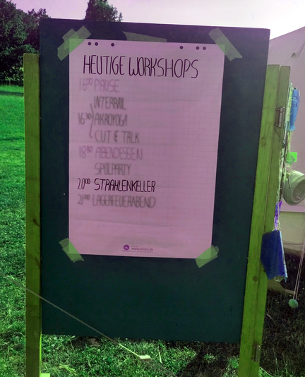 Auf einer saftig-grünen Wiese irgendwo im Nirgendwo steht ein Holzaufsteller mit Flipchart-Plakatbogen, auf dem ein Workshop-Programm gelistet ist.

Programmpunkt für 20:00 Uhr: STRAHLENKELLER