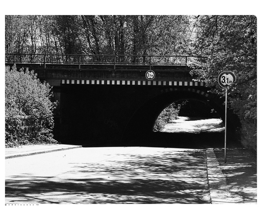 Schwarzweißfotografie: eine breite Straße die unter einer alten Eisenbahnbrücke aus Stein sich verengt, diese hat oberhalb ein Geländer und einen halbrunden dunklen Torbogen, unterhalb sind Warnhinweise auf die Durchfahrtshöhe von 3,10 m angebracht, ebenso das Verkehrszeichen rechts im Bild, ringsherum Bäume und Sträucher