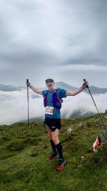 A man in running gear, with his arms outstretched holding a set of running poles.  He has a silly look on his face.

He is stood on grassy ground.  In the background is a cloud inversion and the tips of some Welsh mountains