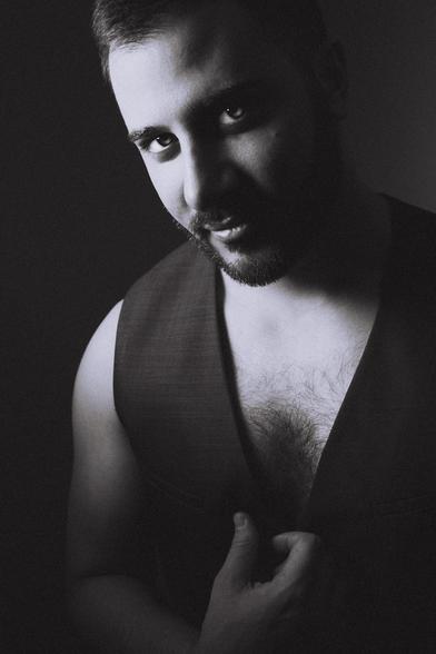 portrait of Turkish American model, shirtless, in a black waist coat, on hand slightly grasping the fabric lining of the vest, shot in stark lighting against a dark background