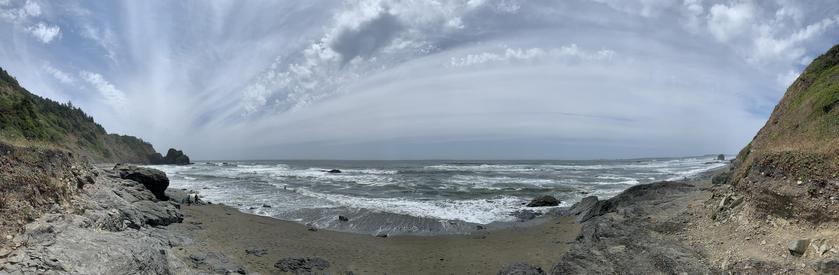 A photograph of a rocky beach, looking out over the water. The format of the photo makes the sky appear curved.