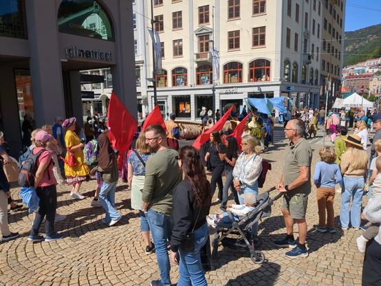 The image captures a bustling scene in Bergen, Norway. A diverse crowd has gathered on the cobblestone pavement, with several individuals holding red flags. In the background, one can see the storefronts of Ginatricot and Norli, along with the traditional buildings characteristic of the city. Tents are scattered about.