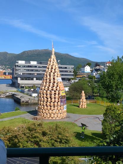 A large wooden structure, resembling a cone or a stylized tree, is situated in a park-like setting, surrounded by greenery and buildings.
