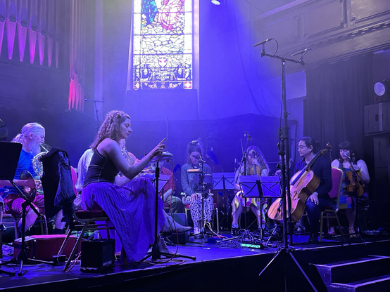 Members of the Glasgow Improvisers Orchestra on an array of instruments (French horn, theremin, saxophone, electronics, double bass, violins, cello) on the stage of St Luke’s in the east end of Glasgow. The stage is bathed in blue-ish light and in the background is a stained glass window with the pipes of a large church organ to the back left.