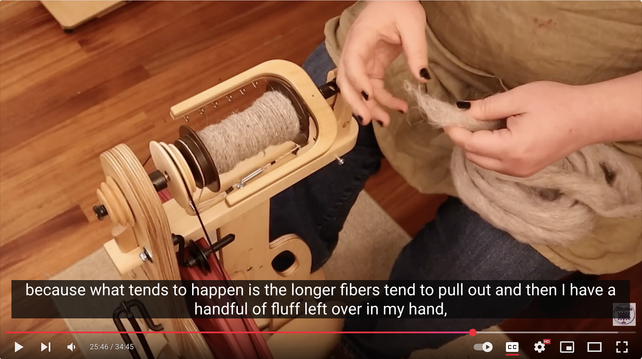 Screenshot from linked YouTube video showing the host spinning a beige wool roving into yarn. It is paused at 25:46 with caption text visible, reading, "because what tends to happen is the longer fibers tend to pull out and then I have a handful of fluff left over in my hand."