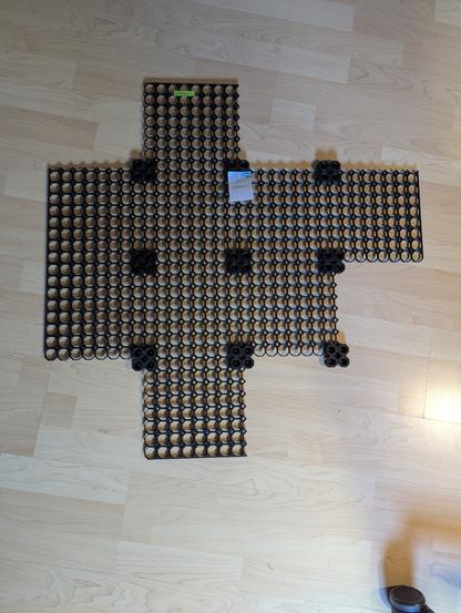 Ein Foto einiger schwarzer Multiboard-Platten, die auf dem hellen Laminat-Boden liegen. Die Platten sind jeweils 20 x 20cm groß und sind an den Ecken mittels Halterungen miteinander verbunden. Die Platten haben große, 8-Eckige Löcher, drumherum jeweils 4 kleinere Löcher. Zu sehen sind 4 Mittelplatten, links davon zwei Platten für die linke Seite, und jeweils eine Platte für den oberen, unteren und rechten Rand.