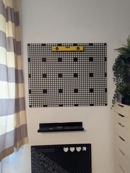 Ein Foto des fertigen Multiboards am einer Wand. Das Board besteht aus 5 x 4 schwarzen Platten und ist 1 Meter breit und 80cm hoch. Oben in der Mitte des Boards habe ich eine Wasserwage festgeklemmt, damit ich beim Einzeichnen der Bohrlöcher sehen konnte, dass es waagerecht hängt.

Unter dem Board ist noch eine schwarze Ablage und ein schwarzen  Skadis-Board von Ikea zu sehen. Rechts daneben ein heller Schrank mit Pflanzen drauf, links ein Vorhang.