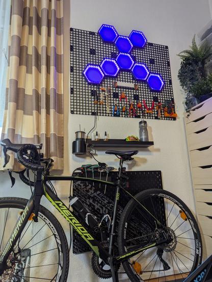 Ein Foto der Wand mit dem Multiboard, vom Boden des Zimmers aufgenommen. Im unteren Bereich sieht man mein Rennrad, darüber eine schwarze Ablage und dann das Multiboard. Inzwischen hängen blau beleuchtete Hexfelder daran, sowie diverses Werkzeug.