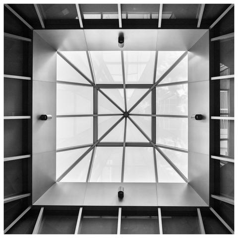 An upward view of a modern atrium featuring a large, square skylight with a geometric grid pattern formed by intersecting metal beams. The beams create smaller squares and triangles, allowing light to pass through. The surrounding walls have shelves, and small lights are evenly spaced around the space, contributing to a sense of depth and openness.