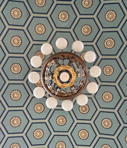 Blaugraue, schwarz umrandeteSechsecke mit einem beigefarbenen Blumenmotiv in der Mitte, zentral eine von unten aufgenommene Deckenlampe aus vielen runden, kugelförmigen Lampen im Kreis angeordnet und zentral auch einem blütenförmigen Lampenschirm