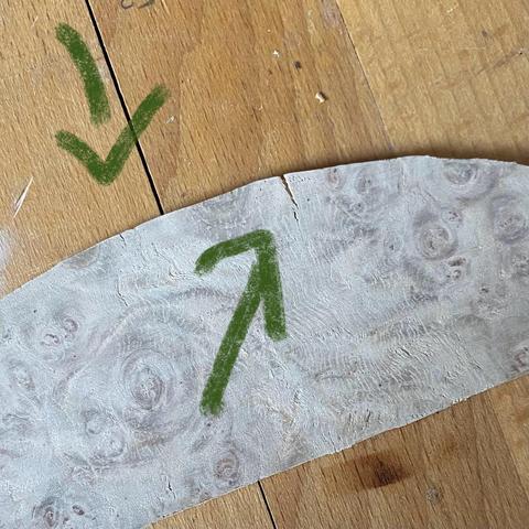 A piece of wood on top of which sits a curved piece of modelling wood with veneer glued on which has contracted after gluing and sanding cracked. Two green arrows point to the cracks.