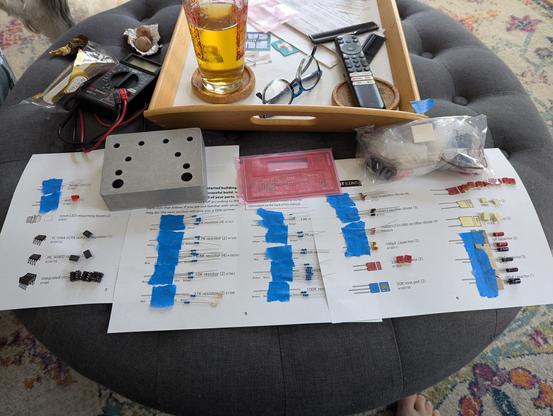 An ottoman covered in sheets of paper with electronics components taped to them, a red PCB, and an unpainted pedal case. A multimeter, glass of beer, and tv remote in the background.