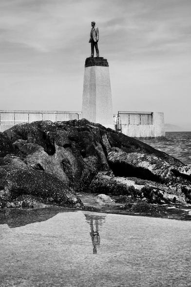 A monochrome photograph features the bronze statue of Roger Casement standing atop a tall, tapered stone plinth on a rocky outcrop in Dun Laoghaire. The statue is reflected in a calm pool of water in the foreground, with the sea and a hazy coastline in the background under a mostly clear sky.