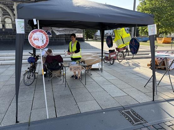 Ein Pavillon mit blauem Dach, rechts und links hängen Zettel und vorne steht ein in Plastik eingeschweisstes Verkehrsschild "Zweiradfahrer dürfen von PKWs, LKWs und Bussen nicht überholt werden".