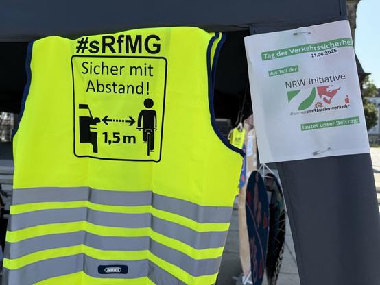 Links eine neongelbe Warnweste mit dem Hashtag #sRfMG und dem Text Sicher mit Abstand sowie ein PKW und ein Radfahrer zwischen denen 1,5 m Abstandspfeil aufgedruckt ist. Rechts das Logo der NRW Initiative #sicherimStraßenverkehr.
