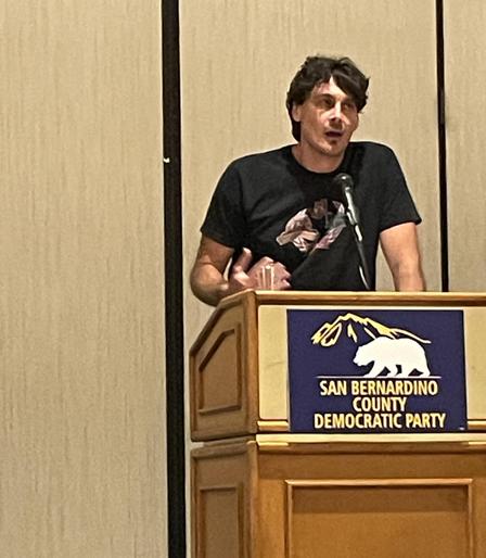 Chris Kluwe behind a podium with the San Bernardino County Democratic Party logo.