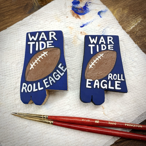 Two little 2-inch-tall pieces of wood cut into the shape of the state of Alabama, painted navy blue, with a football in the center diagonally. Above the football it says “WAR TIDE” and below the football it says, “ROLL EAGLE.”