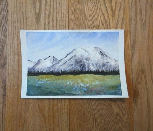 This is a watercolor landscape painting of Mount Rainier. It is in landscape orientation. The painting is roughly divided in thirds horizontally: top third sky, middle third mountain range, bottom third meadow. The sky swoops in Ultramarine Blue with lighter values just above the mountain tops. The white capped mountains have shadowing to define its shape and is dotted with trees here and there. A dark tree line is at its base. The meadow is yellow green in the distance and gets darker moving to the foreground with splattering of blue and purple flowers.