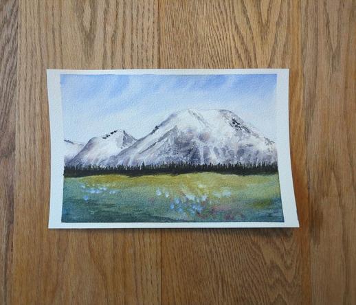 This is a watercolor landscape painting of Mount Rainier. It is in landscape orientation. The painting is roughly divided in thirds horizontally: top third sky, middle third mountain range, bottom third meadow. The sky swoops in Ultramarine Blue with lighter values just above the mountain tops. The white capped mountains have shadowing to define its shape and is dotted with trees here and there. A dark tree line is at its base. The meadow is yellow green in the distance and gets darker moving to the foreground with splattering of blue and purple flowers.