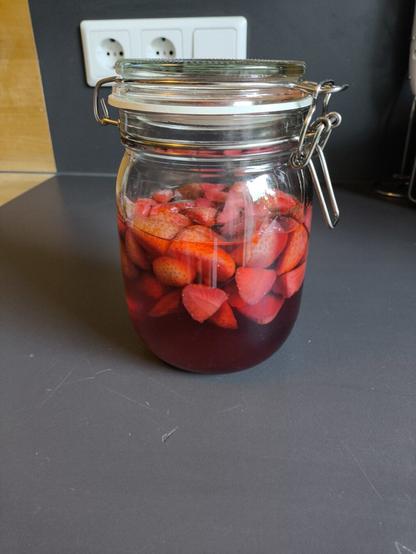 Ein großes Einmach-Glas mit roter Flüssigkeit und geschnittenen Erdbeeren darin.