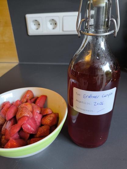 Links eine Schüssel mit geschnittenen Erdbeeren. Rechts ist eine Flasche mit roter Flüssigkeit darin zu sehen. Auf der Flasche ist ein Etikett auf dem "Erdbeer Campari" steht.