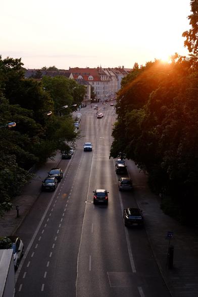 A wide street in a quiet urban neighborhood during sunset, lined with parked cars and flanked by tall green trees. A few vehicles drive down the road, and the warm orange sun peeks through the trees, casting a golden glow on the scene. Residential buildings with red rooftops are visible in the distance.
