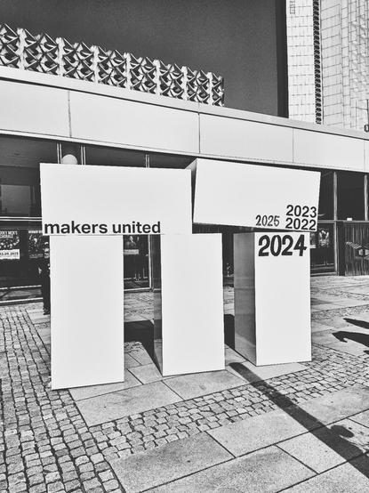 Das Bild zeigt drei weiße, rechteckige Säulen, die auf einem gepflasterten Gehweg stehen. Auf den Säulen sind verschiedene Texte zu sehen. Die linke Säule trägt den Text "makers united". Die mittlere Säule ist leer, während die rechte Säule die Jahre "2023", "2024", "2025" und "2022" in großen, schwarzen Ziffern zeigt. Die Säulen stehen vor einem Gebäude mit einem modernen, geometrischen Muster an der Fassade. Der Hintergrund zeigt ein Teil eines weiteren Gebäudes, das hoch und mit Glasfassade ausgestattet ist. Die gesamte Szene ist in Schwarz-Weiß fotografiert, was die Kontraste und die Textur der Säulen und des Gehwegs hervorhebt.