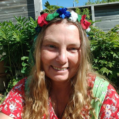 Head and shoulders of a white middle aged woman with long light brown hair. She is wearing a wreath with green leaves and blue, white, and red flowers, all made from fabric. She has a red blouse and a light green dirndl style dress.
