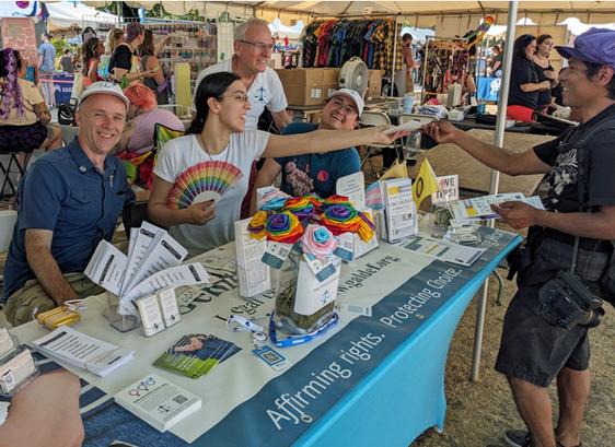GALDEF's 2024 Portland Pride table.  Pride roses in the center, Allie handing out a flier while holding a rainbow fan.  Tim Hammond of GALDEF standing behind, and Perry of GAC sitting behind.