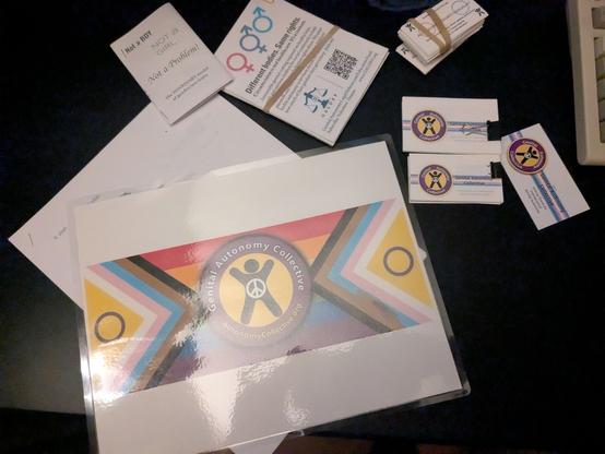 Black desktop with various printed GAC and GALDEF materials.   A laminated letter page sized banner with a mirrored progress flag and GAC logo.