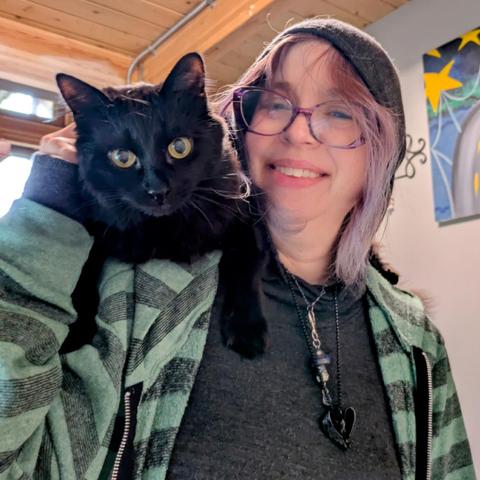 A neglected black cat calls put for attention from his purple haired human's shoulder