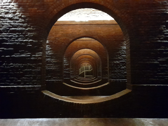 The inside of the old water tanks of Brno. A symmetrical view of an opening in a dark brick wall, through which another similar wall with another opening can be seen. Through this opening six more walls with openings are visible, each time smaller as it is further away. Through the last opening a staircase is visible going up to the right.