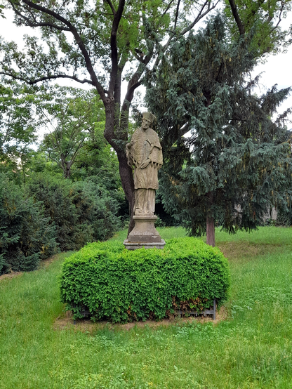 A statue of a saint holding a cross in a park. Behind the statue are some trees and around it are neatly trimmed bushes.