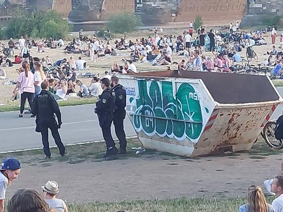 Drei Polizisten stehen neben einem Müllcontainer mit Grafitti