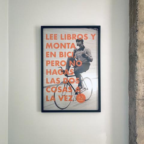 Una fotografía de un cuadro. En él se ve a una persona montando en bicicleta, bebiendo café y leyendo un libro. Sobre el cuadro se puede leer: “Lee libros y monta en bici, pero no hagas las dos cosas a la vez”.
