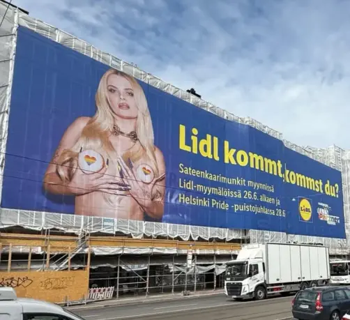 Lidlin valtavankokoinen mainoslakana rakennustelineissä. Sinisessä lakanassa on keltaista tekstiä ja kuva yläosattomasta Erika Vikmanista, joka peittää rintansa kahdella sateenkarisydämellä kuvioidulla munkilla.