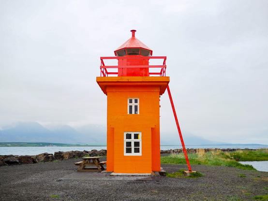 A moody photo of a fairly short, orange lighthouse with a red top, a ladder on the right hand side that leads to the top, and a picnic bench on the left hand side. A fjord and foggy mountains are in the background.