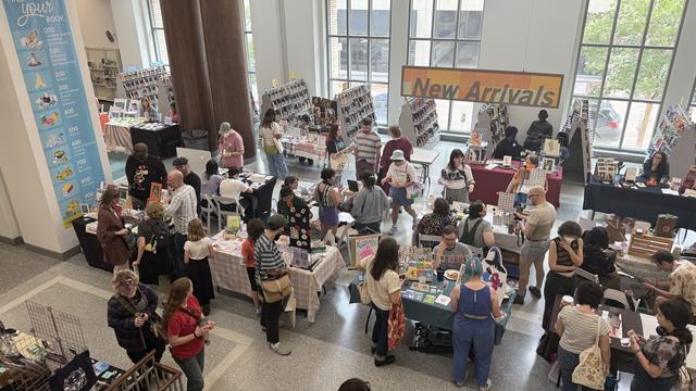 A bustling event scene inside a public library featuring a crowd of people browsing tables filled with merchandise, including books and art. There are displays showcasing "New Arrivals" and various items for sale.