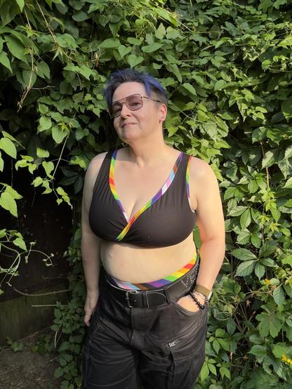 A person stands in front of lush green foliage, wearing a black bikini top with a rainbow-colored accent and matching bikini bottoms. They have short, blue hair and are wearing sunglasses, along with black cargo shorts with belt and accessories like bracelets.