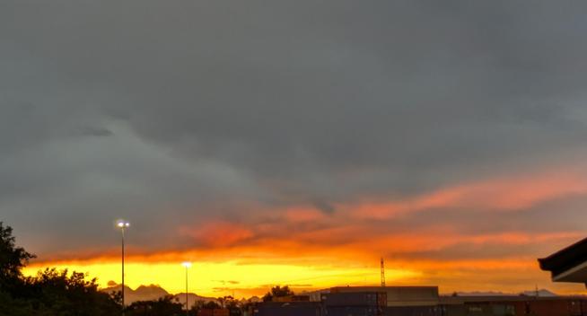 cielo dominato da nuvole scure e grigie nella parte superiore, che contrastano nettamente con una fascia vibrante di colori caldi all'orizzonte. Questa fascia mostra intense tonalità di arancione brillante e giallo dorato, suggerendo la presenza del sole appena tramontato, e sullo sfondo le vette del Monte Rosa