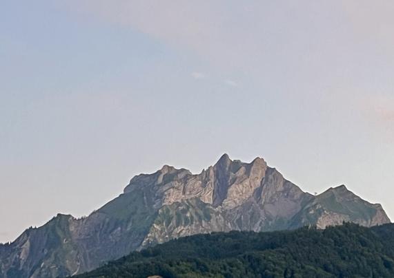 Pilatusberg nach Sonnenuntergang. Die bewaldeten Hügel vor dem Berg sind Sattgrün. Der Himmel ist mit Schleierwolken bedeckt und erscheint gräulich.