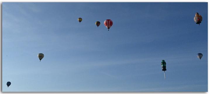 Acht Heißluftballons fahren am Himmel, verteilt in verschiedenen Höhen und Abständen. Der Himmel ist wolkenlos und blau. Die Ballons haben unterschiedliche Farben und Formen. Einige Ballons sind rund und bunt, einer ist rot-weiß gestreift. Zwei auffällige Ballons haben Sonderformen: Einer sieht aus wie ein Schraubendreher mit schwarz-grünem Griff, unter dem Korb hängt der Schaft. Ein anderer sieht aus wie ein dicker Tankwart.