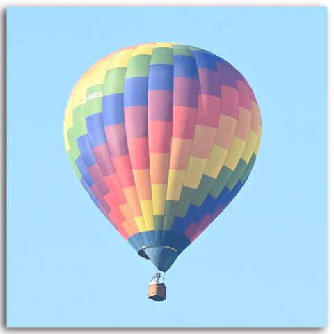 Ein einzelner großer Heißluftballon in Nahaufnahme vor blauem Himmel. Der Ballon hat ein Muster aus bunten, rechteckigen Farbfeldern in Regenbogenfarben – darunter Gelb, Grün, Blau, Rot und Violett.